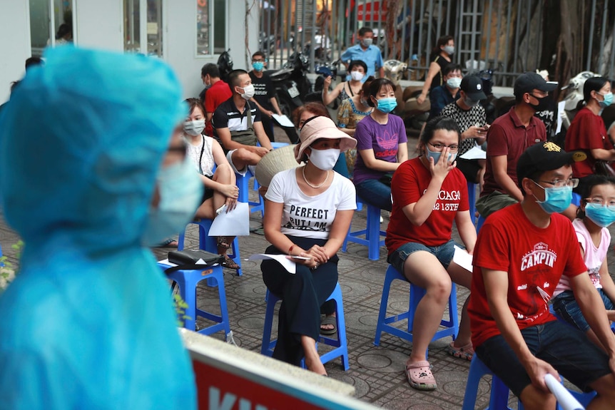People in face masks sit waiting on plastic stools outdoors.