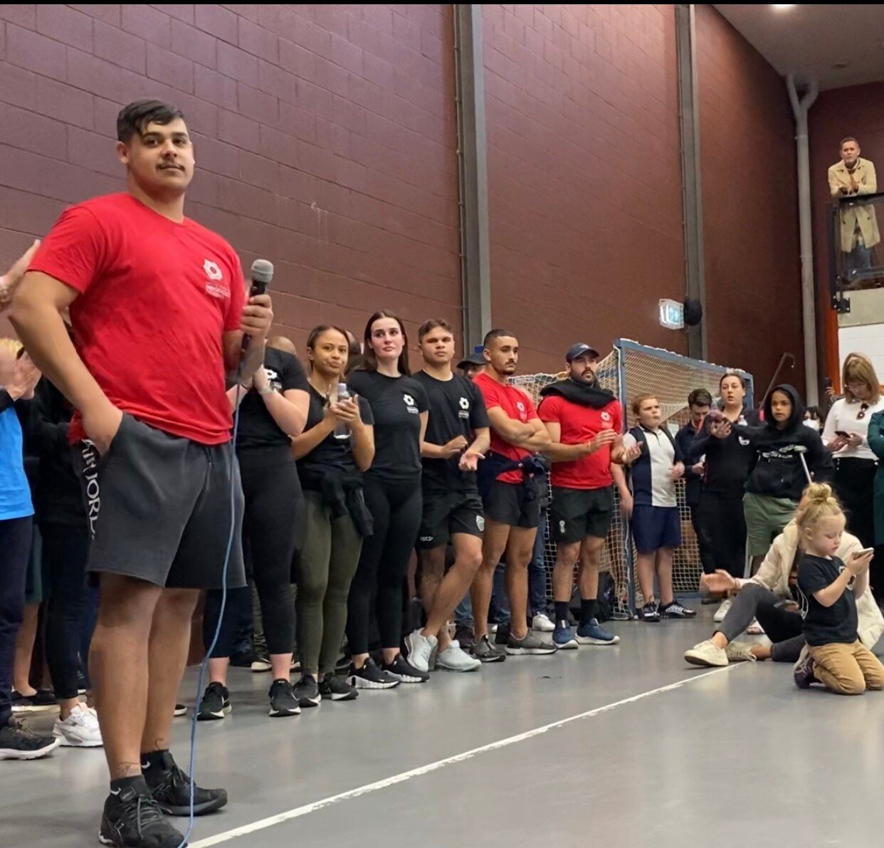 A man holds a microphone as he speaks to people gathered on an indoor basketball court.