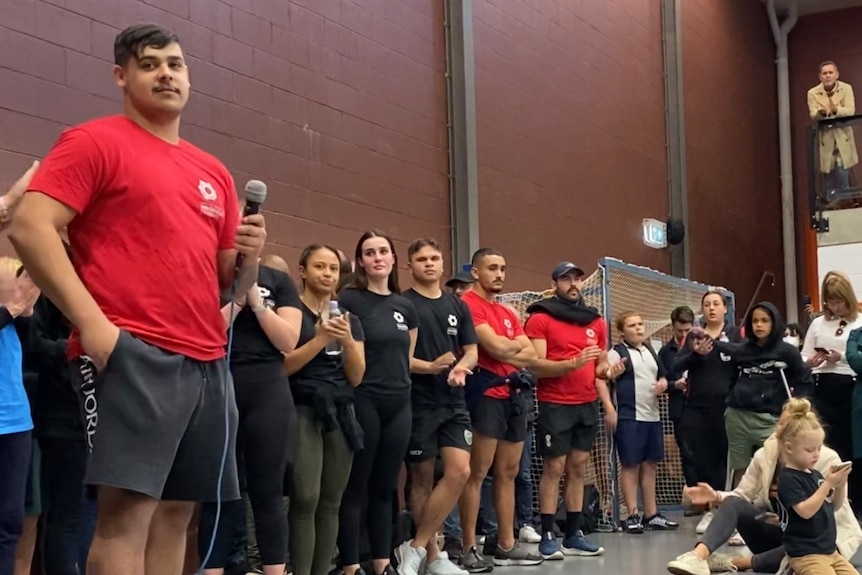 A man holds a microphone as he speaks to people gathered on an indoor basketball court.