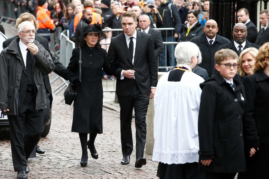 Wide shot of numerous people walking on a road with a crowd behind them.