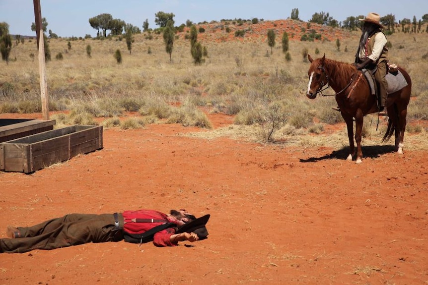 Leslie's character lies dead on red earth while John's character looks on a top a horse, a bush vista in the background.