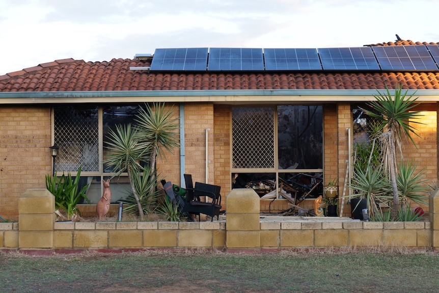 Brick house with solar panels and green plants outside, windows blackened and smashed.