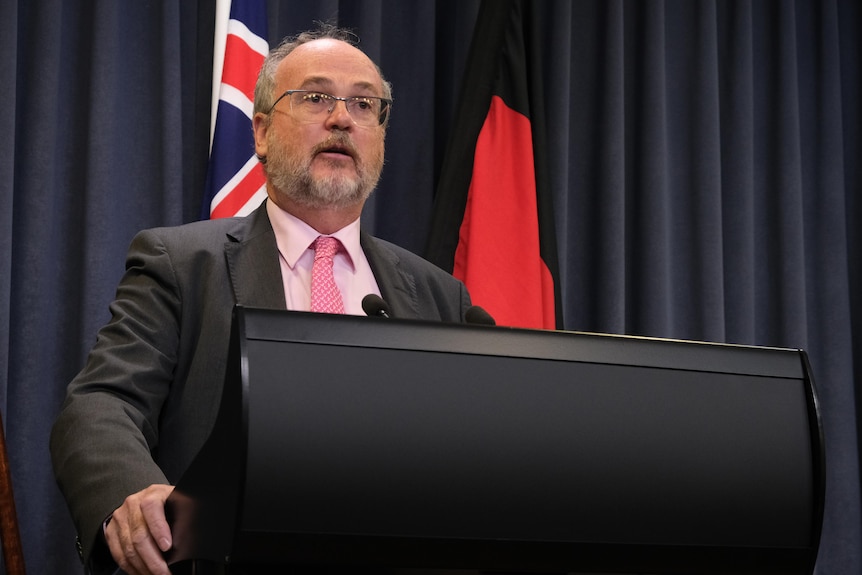 Bill Johnston wears a pink suit and tie with a black suit jacket while speaking at a lecturn