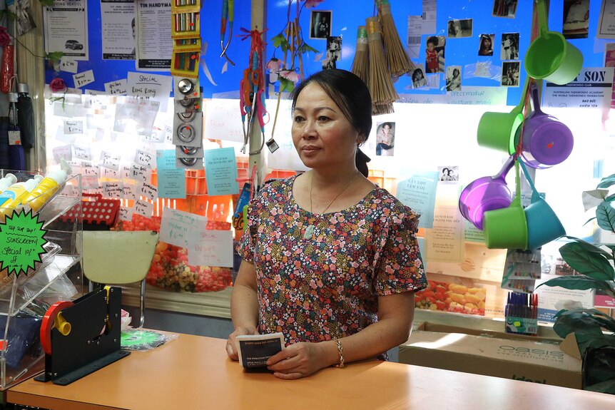Carina Hoang as her character Iris, she is standing behind the counter of her shop, she's wearing a floral dress.
