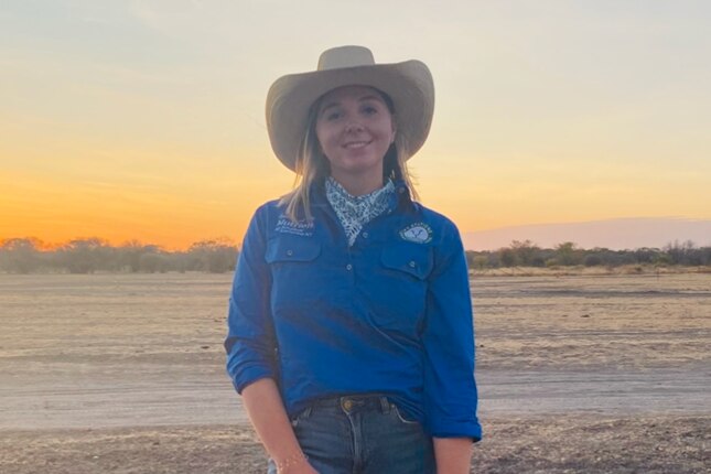 Woman in outback cowgirl wear smiles at camera on cattle station at sunset
