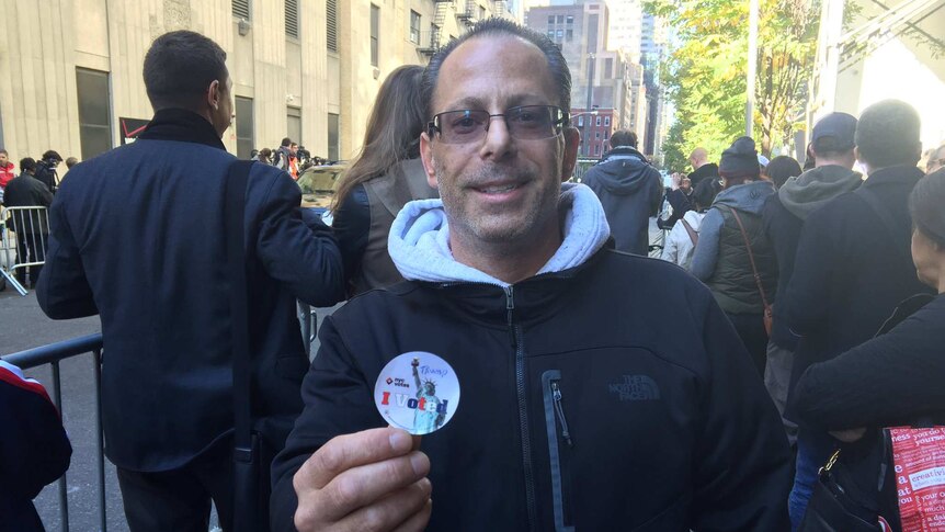 A man in New York holds up an "I voted" sticker with "Trump" handwritten on it.