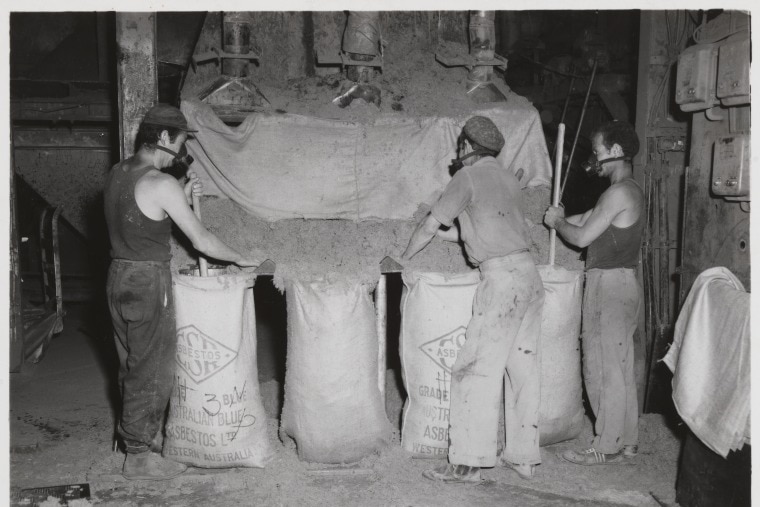 Black and white photo of three men wearing masks and singlets, leaning over table covered in large dark matter.