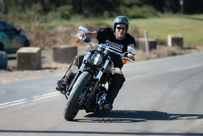 Man wearing a black motorcyle helmet and black t-shirt rides a motorbike on a road.