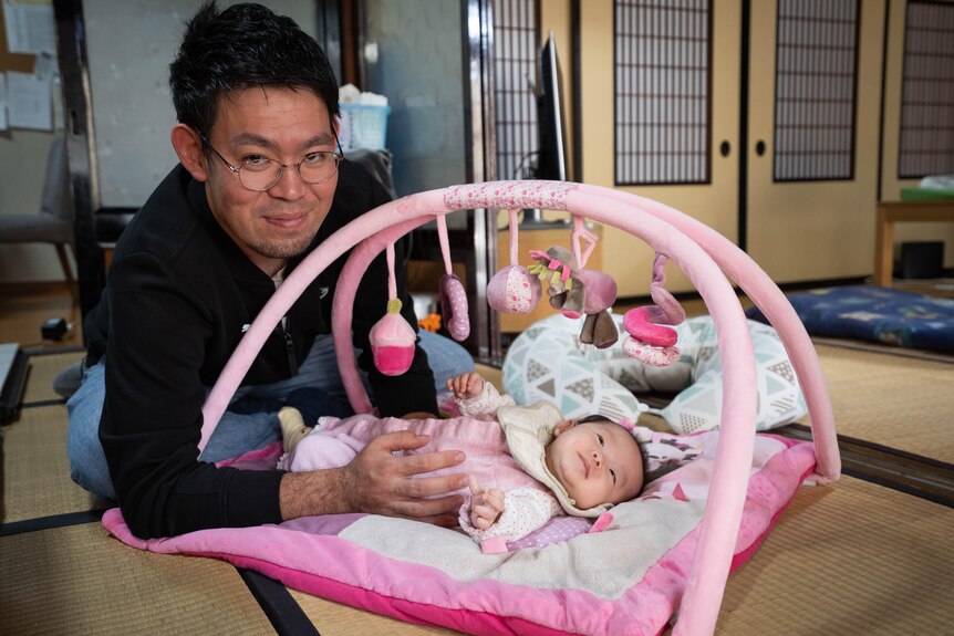 A man dressed in a black shirt lays down holding his baby under a pink mobile.