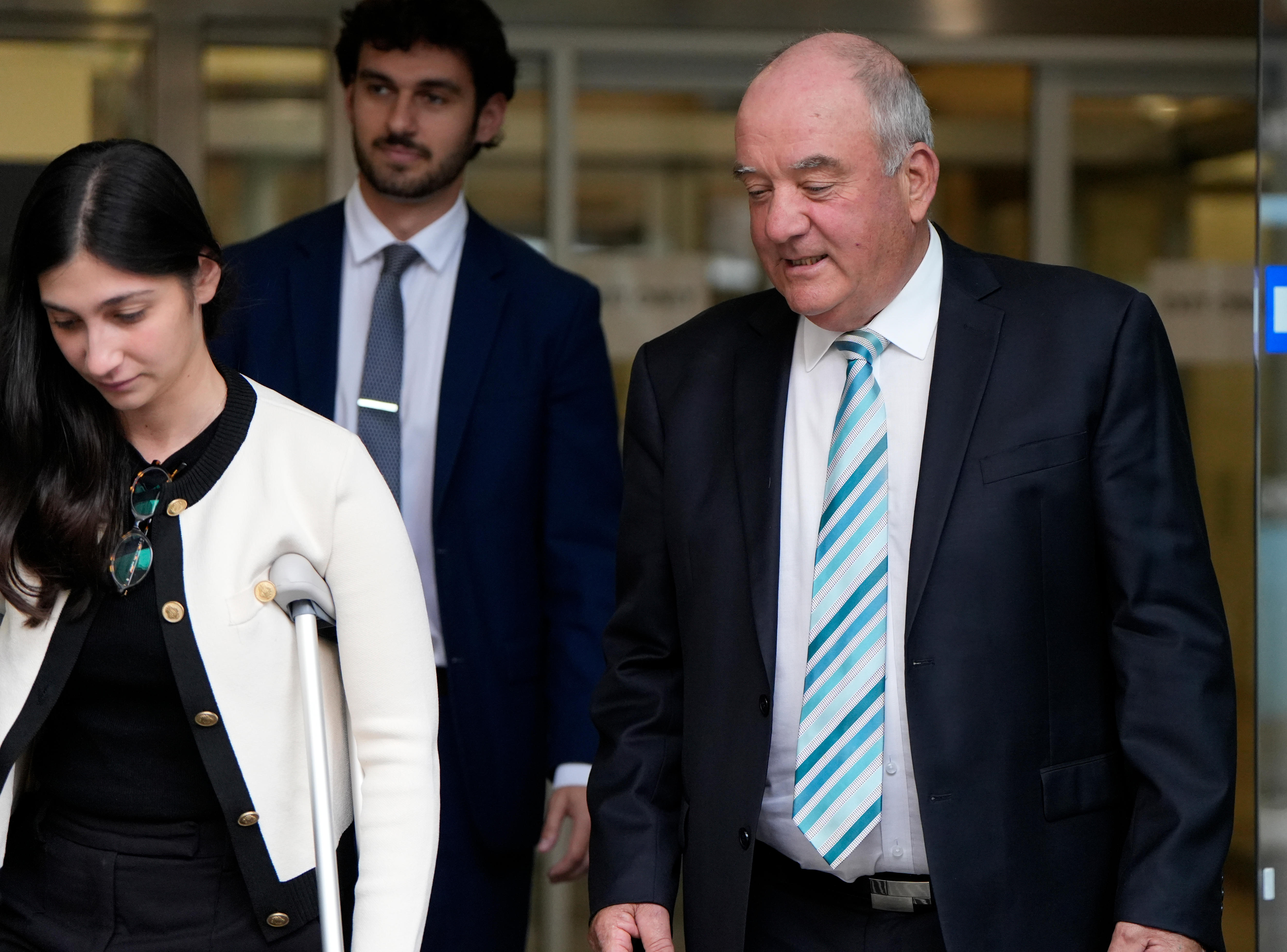 A man in a dark jacket, white shirt and tie leaves a building with a woman and man. 