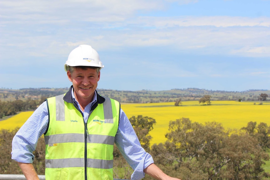 A smiling man wearing a white hard hat with a canola paddock in the background.