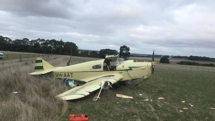 A mostly intact vintage two-seater plane sits in a paddock surrounded by small debris.