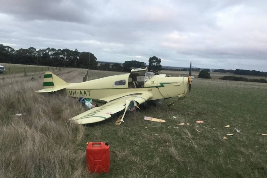A mostly intact vintage two-seater plane sits in a paddock surrounded by small debris.