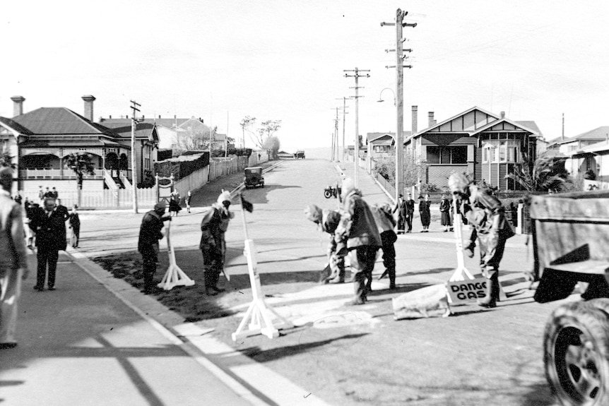 Black and white image of several men in protective suits on a suburban street with a sign reading: Danger gas