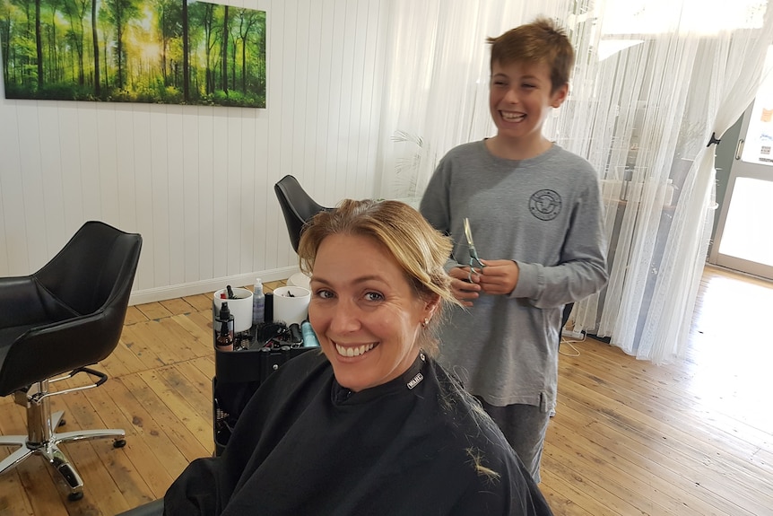 Narelle at the hairdressers with chopped hair smiling, her son Lachlan holding scissors laughing behind her.