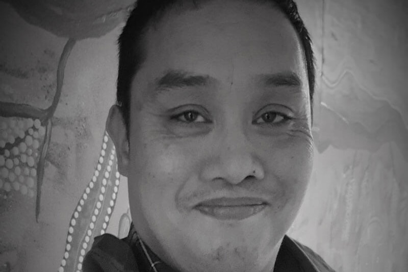 A black and white photo of a smiling man with short black hair sitting in front of an Aboriginal painting.