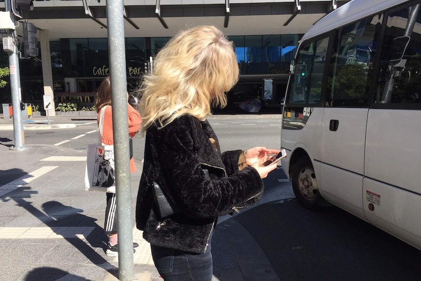 A woman staring at her phone on a street corner as a mini bus crosses her path