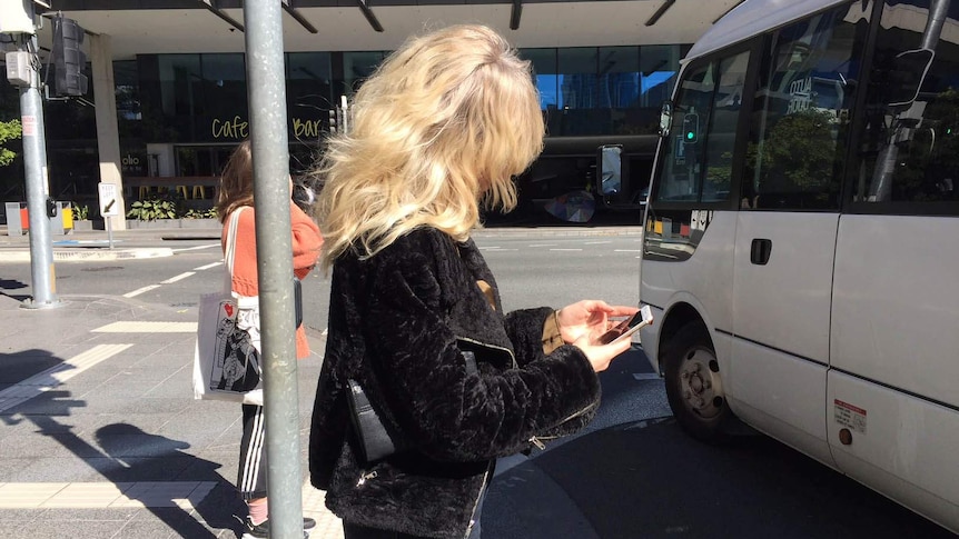 A woman staring at her phone on a street corner as a mini bus crosses her path