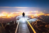 Man stands at the end of boardwalk lookout on a mountain with city lights in background.