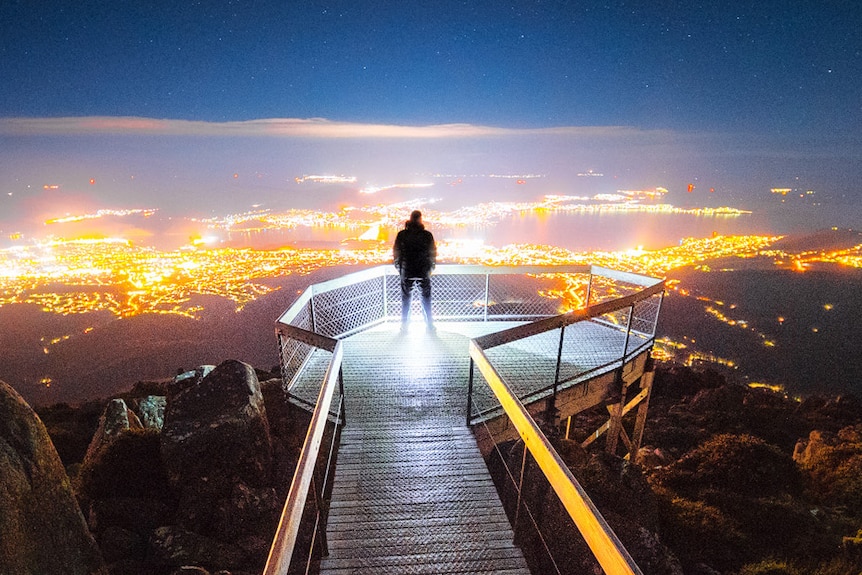 Man stands at the end of boardwalk lookout on a mountain with city lights in background.