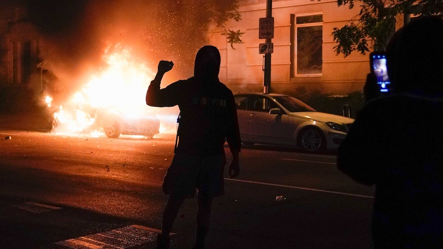 A demonstrator is filmed in front of a burning car during a protest