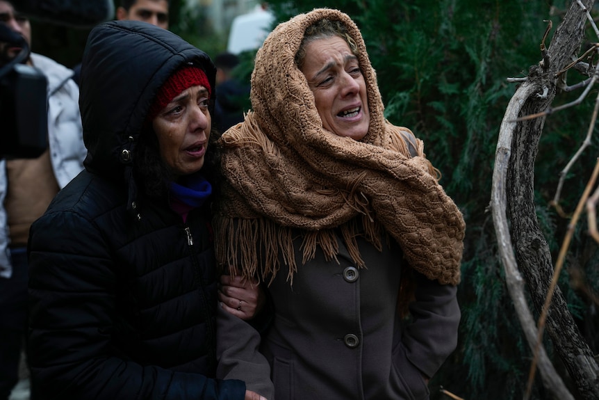 Two women stand together holding eachothers arms crying. 