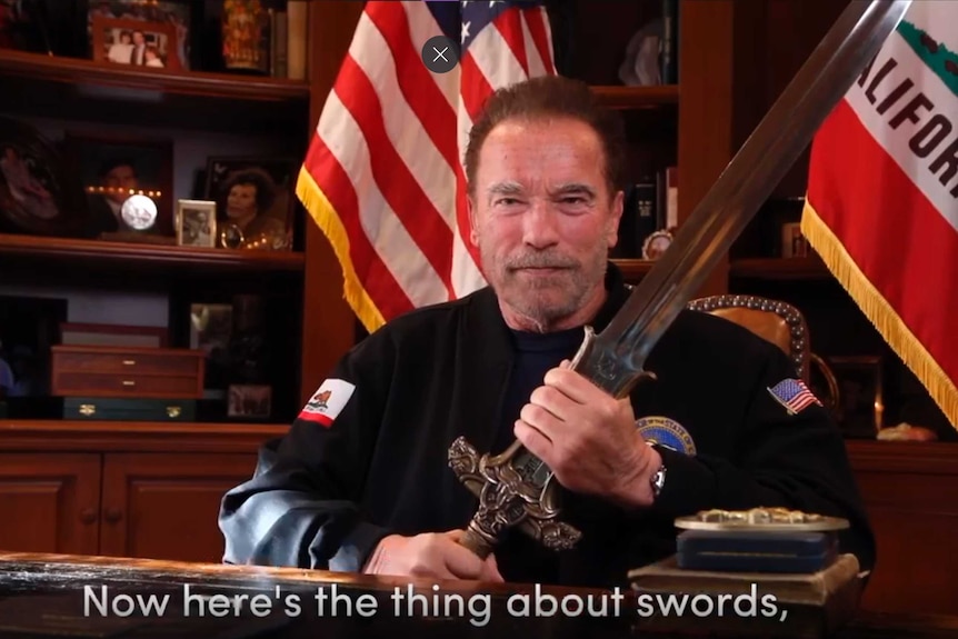 An elderly man holds a sword in two hands at a desk in an office with a California flag behind him.