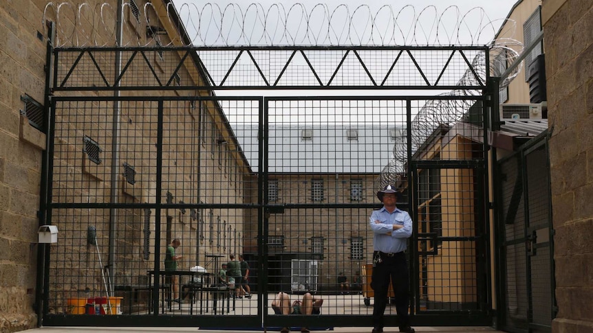 A guard stands in front of a razor wire fence behind which inmates can be seen talking and exercising.