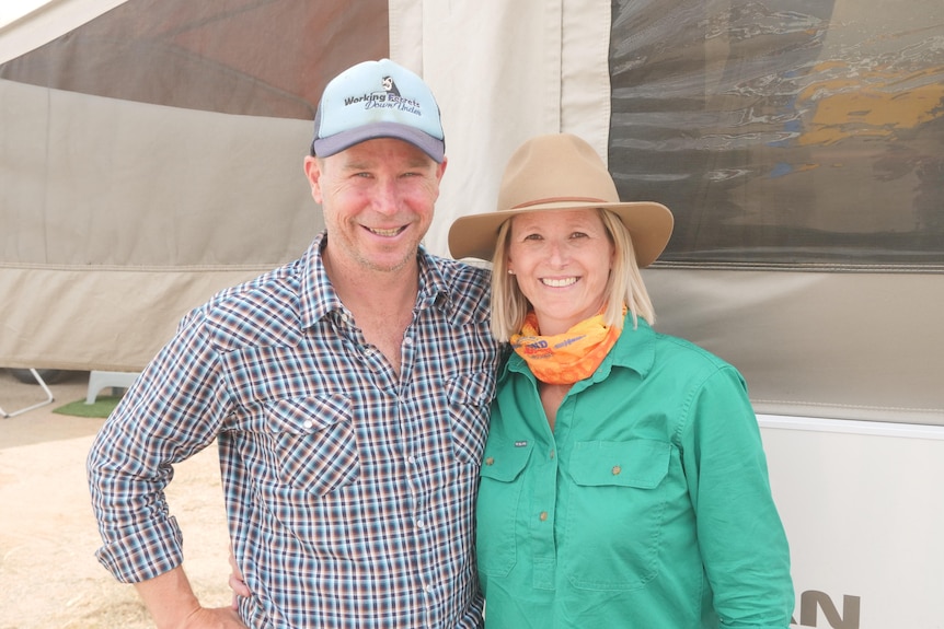 Am man in a checked shirt and cap smiles next to a woman in a green shirt and wide-brimmed hat