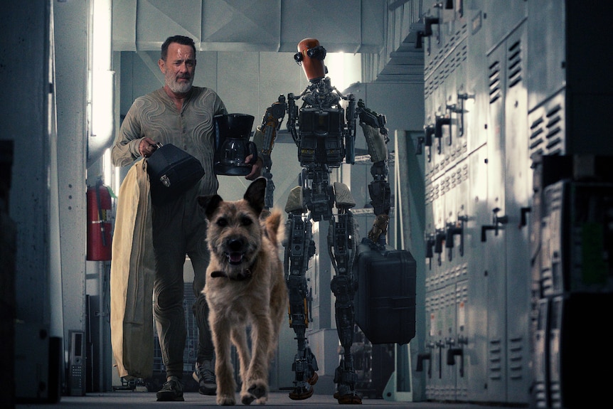 Scruffy bearded middle-aged man in dirty grey shirt carries a kettle and bag beside a metal human-shaped robot and wolfhound.