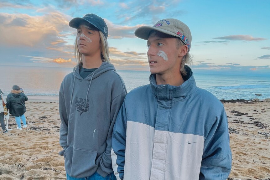 two young men in jumpers and hats standing on the beach with the ocean behind