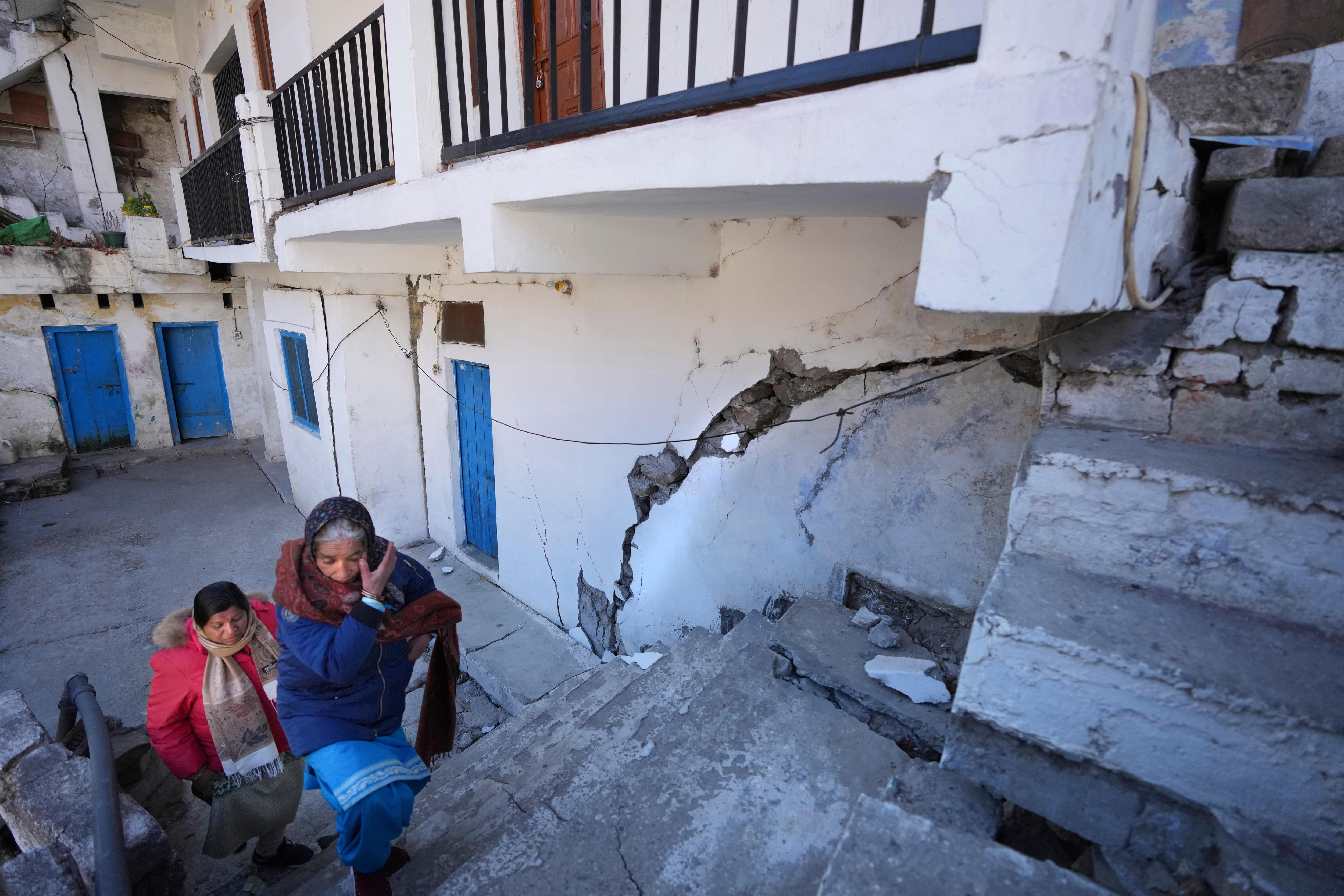 A woman wipes her face as she walks up staps next to a building with a large crack in the wall.