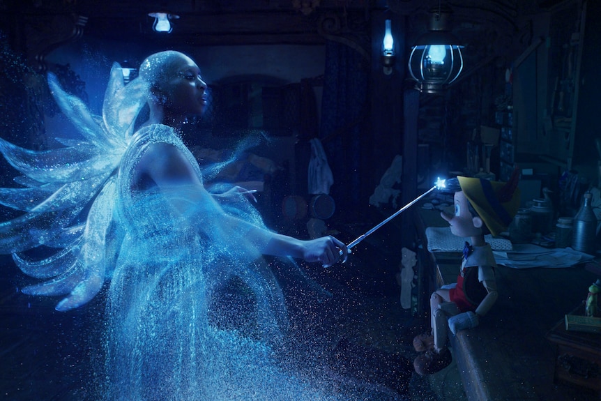 Black woman dressed as magical fairy appears half translucent waving a wand at a small humanoid toy.  