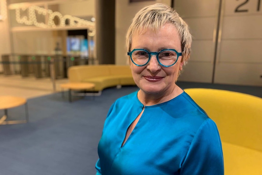 A woman with short blonde hair and blue glasses smiling at the camera.