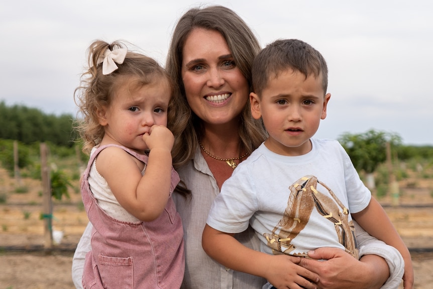 A woman holds two toddlers while smiling and standing on a farm 