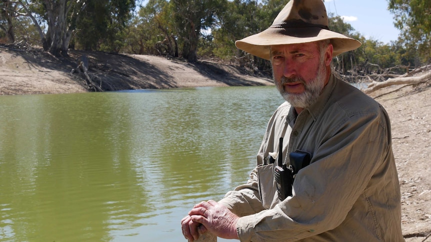 Alan Whyte, wearing a hat, faces the camera with his hands on a depth post on the edge of the Darling River.