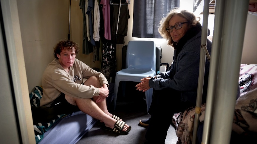 A young man sitting on a mattress and a blonde woman sitting on the edge of a bunk bed in a small room.