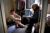 A young man sitting on a mattress and a blonde woman sitting on the edge of a bunk bed in a small room.