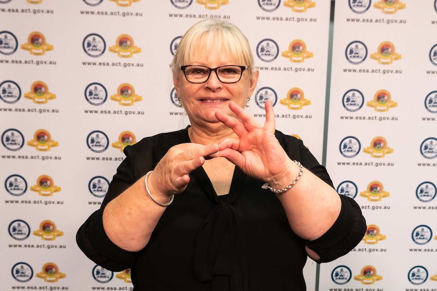 A female Auslan interpreter smiles while signing in front of an ACT Government banner.