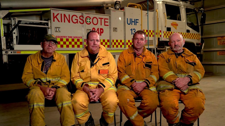 Four male firefighters in yellow protective gear sit in front of a fire truck.