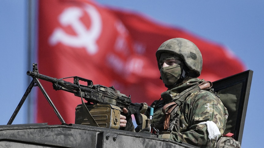 A uniformed soldier looks out of the top of a tank, holding a rifle. A red communism flag billows in the background