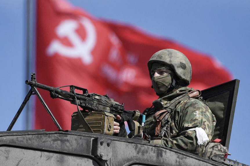 A uniformed soldier looks out of the top of a tank, holding a rifle. A red communism flag billows in the background