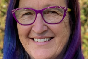 A lady with blue and purple hair with purple glasses smiles at the camera.