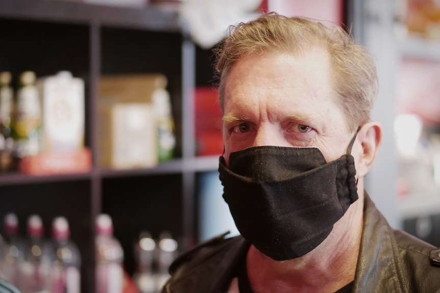 A middle aged man wearing a black mask sits in front of some shelving with food products