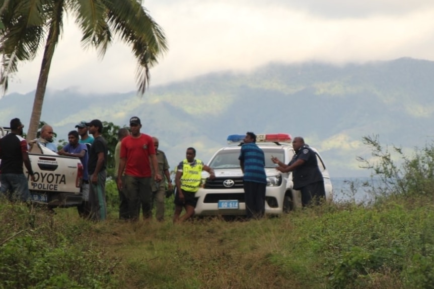 A group of men stand around a police vehicle and truck, with grass in the foreground and mountains in the background.