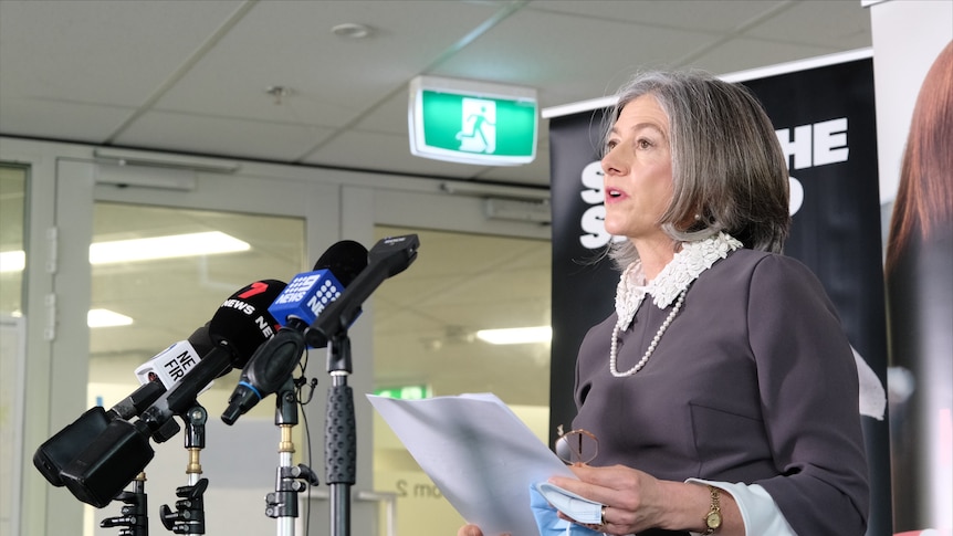 A woman holding a piece of paper and a surgical mask stands in front of media microphones against an SA Health backdrop