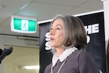 A woman holding a piece of paper and a surgical mask stands in front of media microphones against an SA Health backdrop