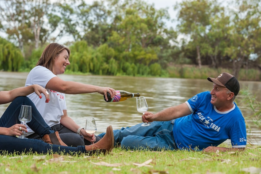 Una mujer blanca sirve una copa de vino espumoso a un hombre blanco mientras se sientan sonrientes junto al río Murray.