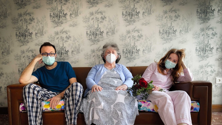 A man, an older lady and a woman all in masks sitting on a couch