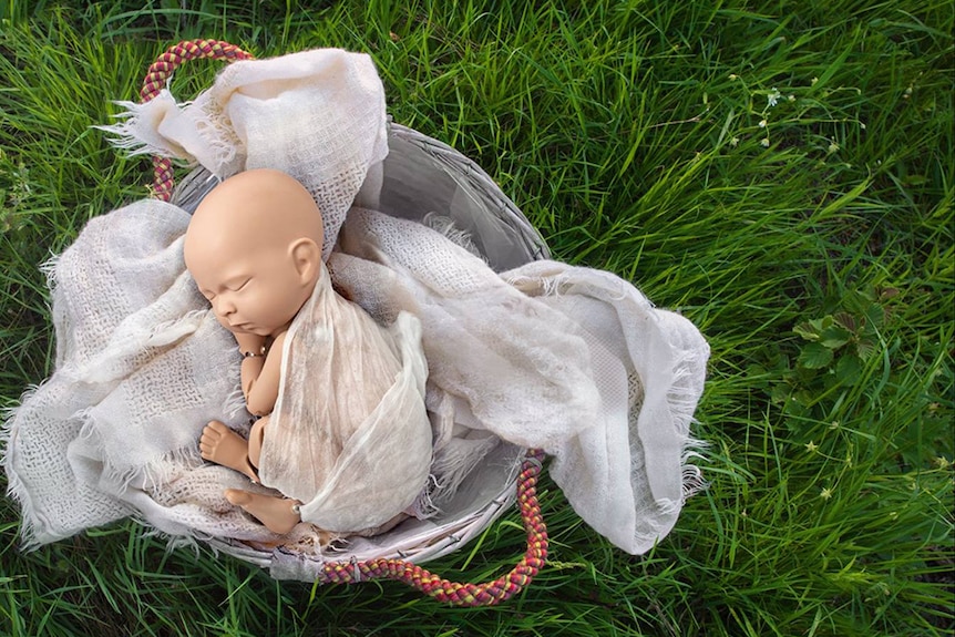A mannequin baby swaddled in white muslin in a basket of blankets on top of green grass.